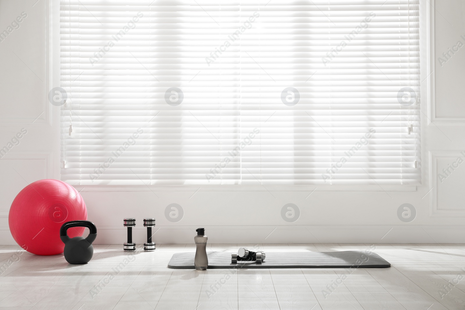 Photo of Exercise mat, dumbbells, kettlebell, fitness ball and bottle near window in spacious room