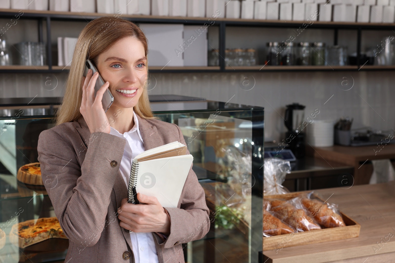 Photo of Happy business owner with notebook and pen talking on phone in bakery shop