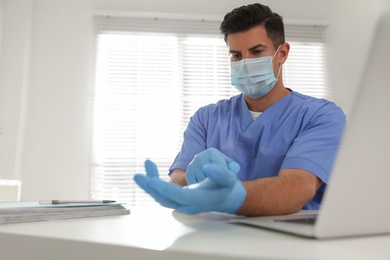 Doctor in protective mask putting on medical gloves at table in office. Space for text