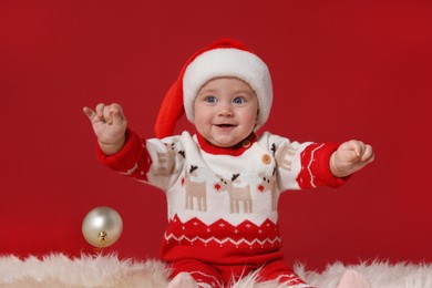 Cute baby in Santa hat with Christmas ball on fluffy carpet against red background