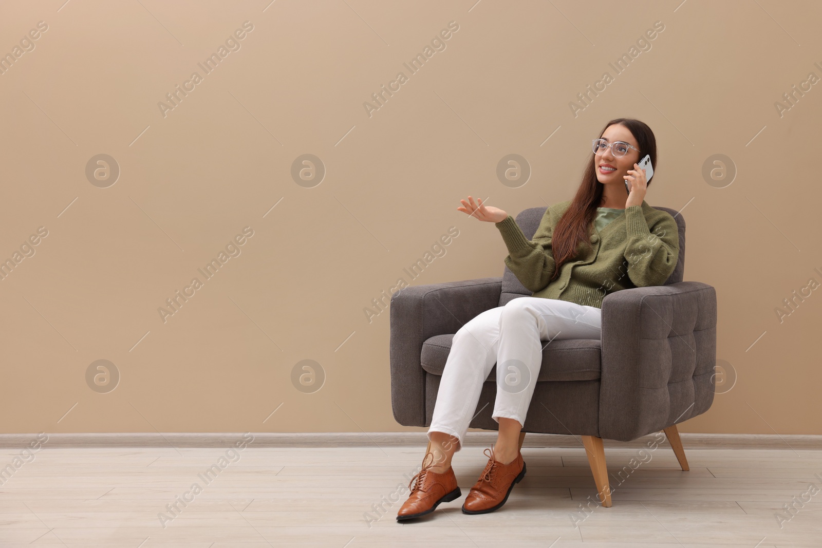 Photo of Beautiful woman talking on smartphone while sitting in armchair near beige wall indoors, space for text