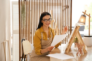 Young woman drawing on easel with pencil at table indoors