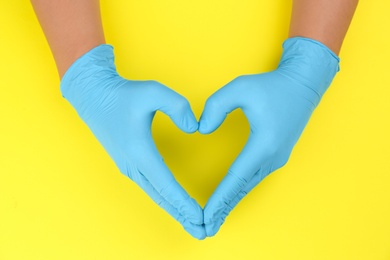 Person in blue latex gloves showing heart gesture against yellow background, closeup on hands