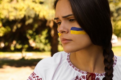 Photo of Sad young woman with drawing of Ukrainian flag on face in park, closeup. Space for text