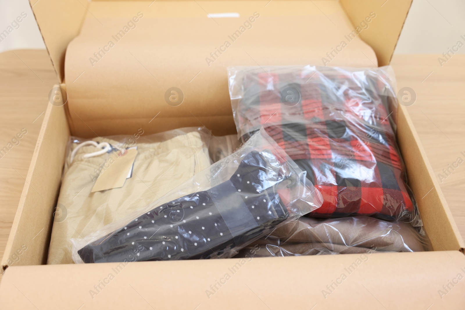 Photo of Open cardboard box with different clothes in plastic bags on wooden table. Delivery service