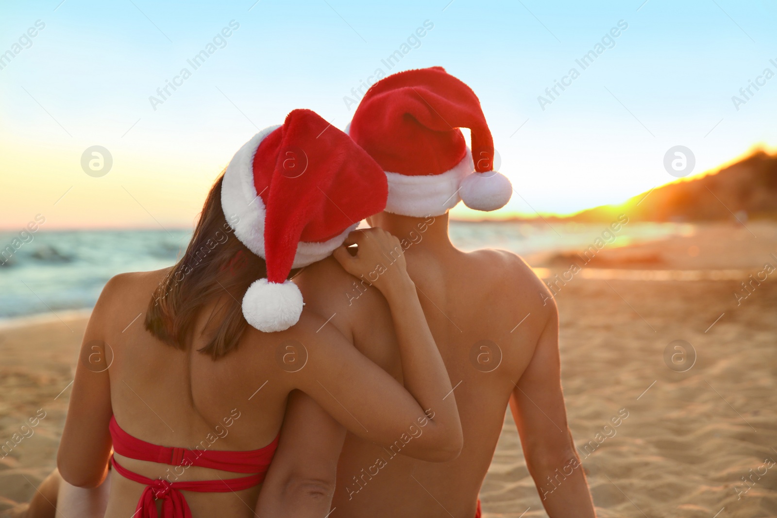 Photo of Lovely couple with Santa hats together on beach at sunset, back view. Christmas vacation