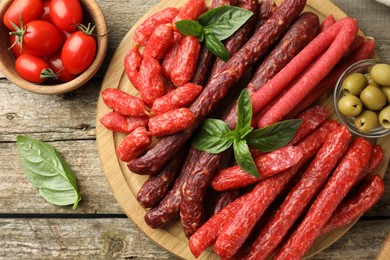 Photo of Different thin dry smoked sausages, basil, tomatoes and olives on wooden table, flat lay