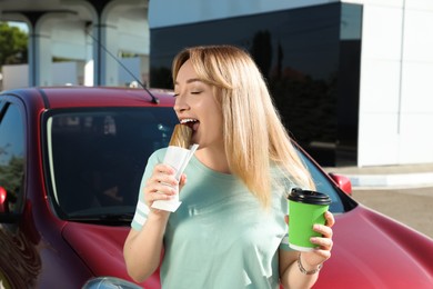 Beautiful young woman with coffee eating hot dog near car at gas station