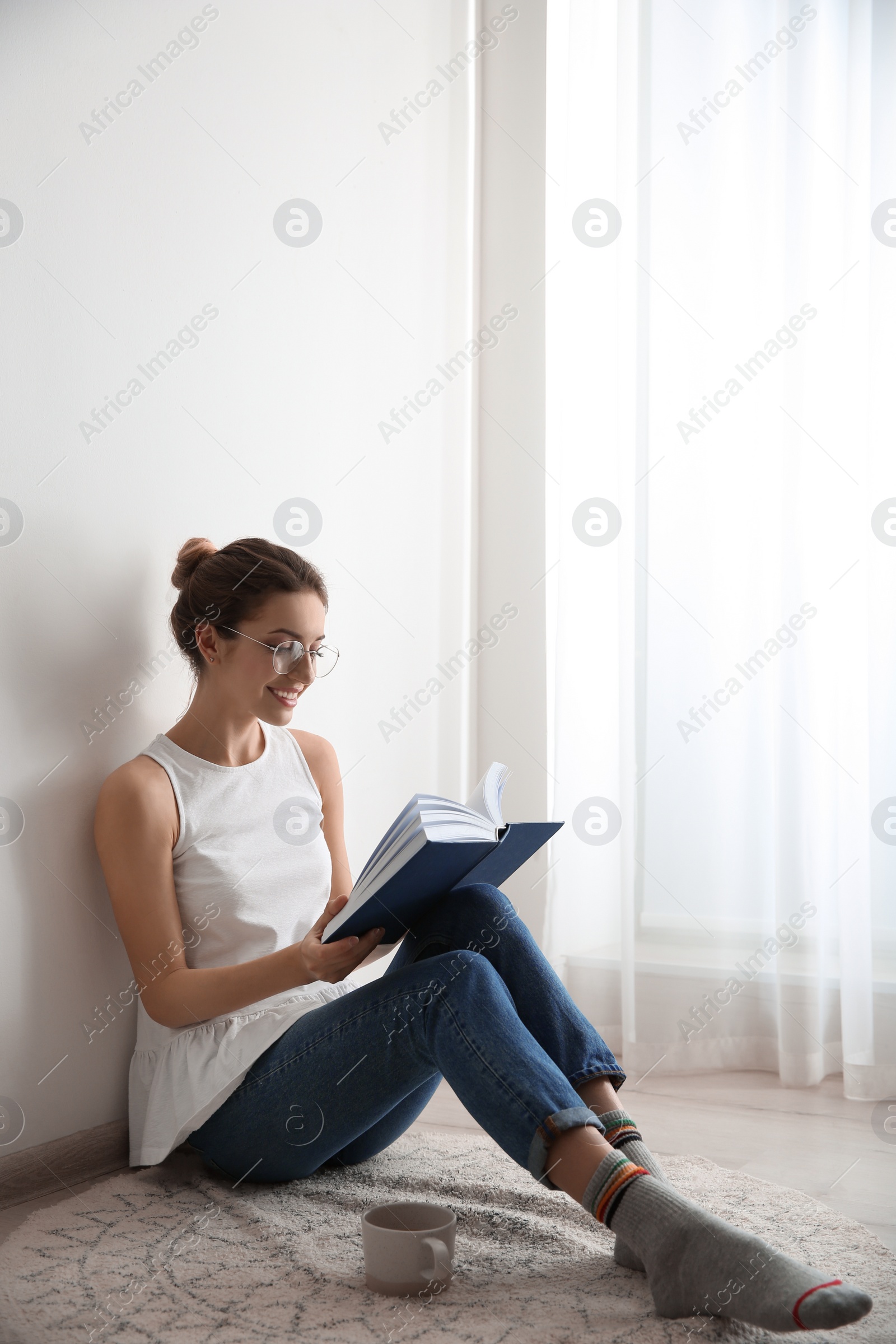 Photo of Young woman with cup of coffee reading book on floor at home