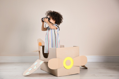 Photo of Cute African American child playing with cardboard plane and binoculars near beige wall