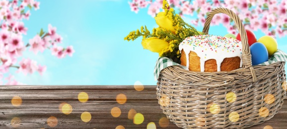 Image of Basket with traditional Easter cake, eggs and flowers on wooden table outdoors, space for text. Banner design