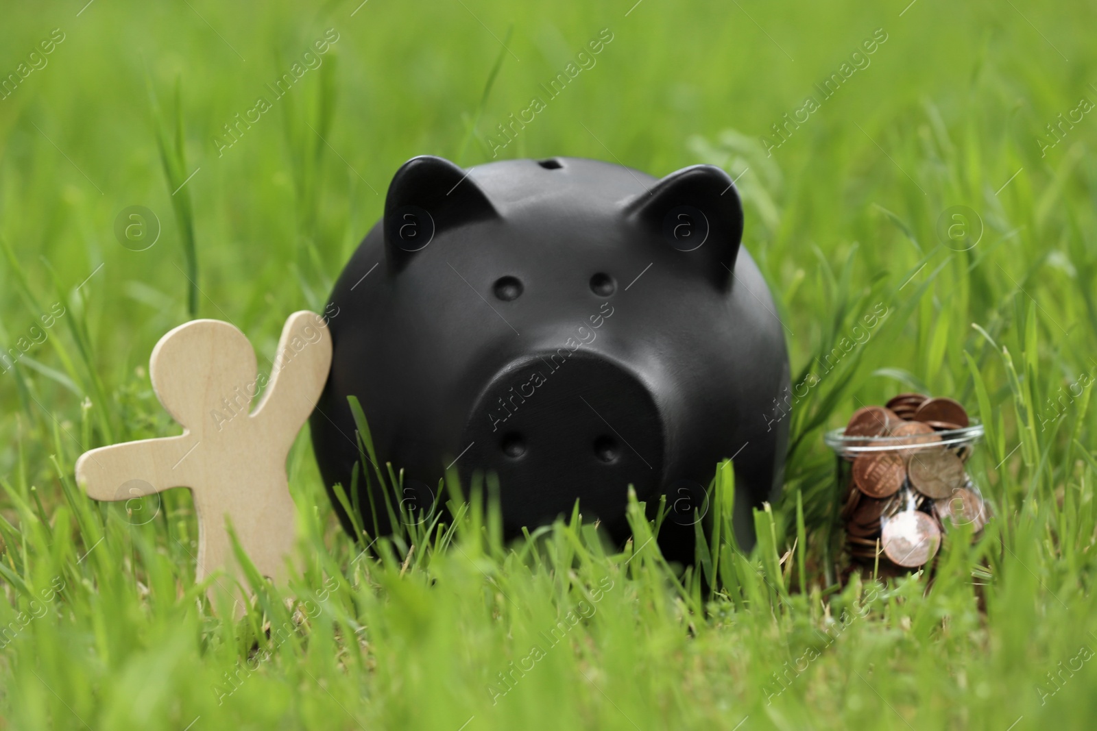 Photo of Cute piggy bank, wooden person and jar with coins on green grass in park