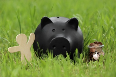 Photo of Cute piggy bank, wooden person and jar with coins on green grass in park