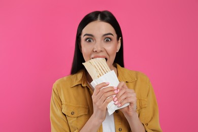 Emotional young woman eating delicious shawarma on pink background