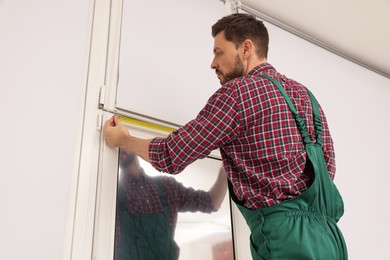 Photo of Worker in uniform using tape measure while installing roller window blind indoors