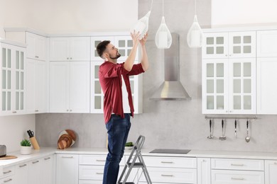 Photo of Young man installing ceiling lamp on stepladder in kitchen