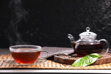 Aromatic hot tea in glass cup, teapot and leaves on table against black background