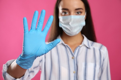 Woman in protective face mask and medical gloves showing stop gesture against pink background, focus on hand