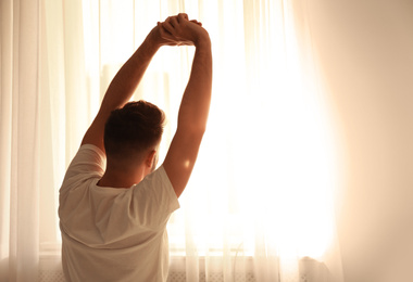 Man stretching near window at home, view from back. Lazy morning