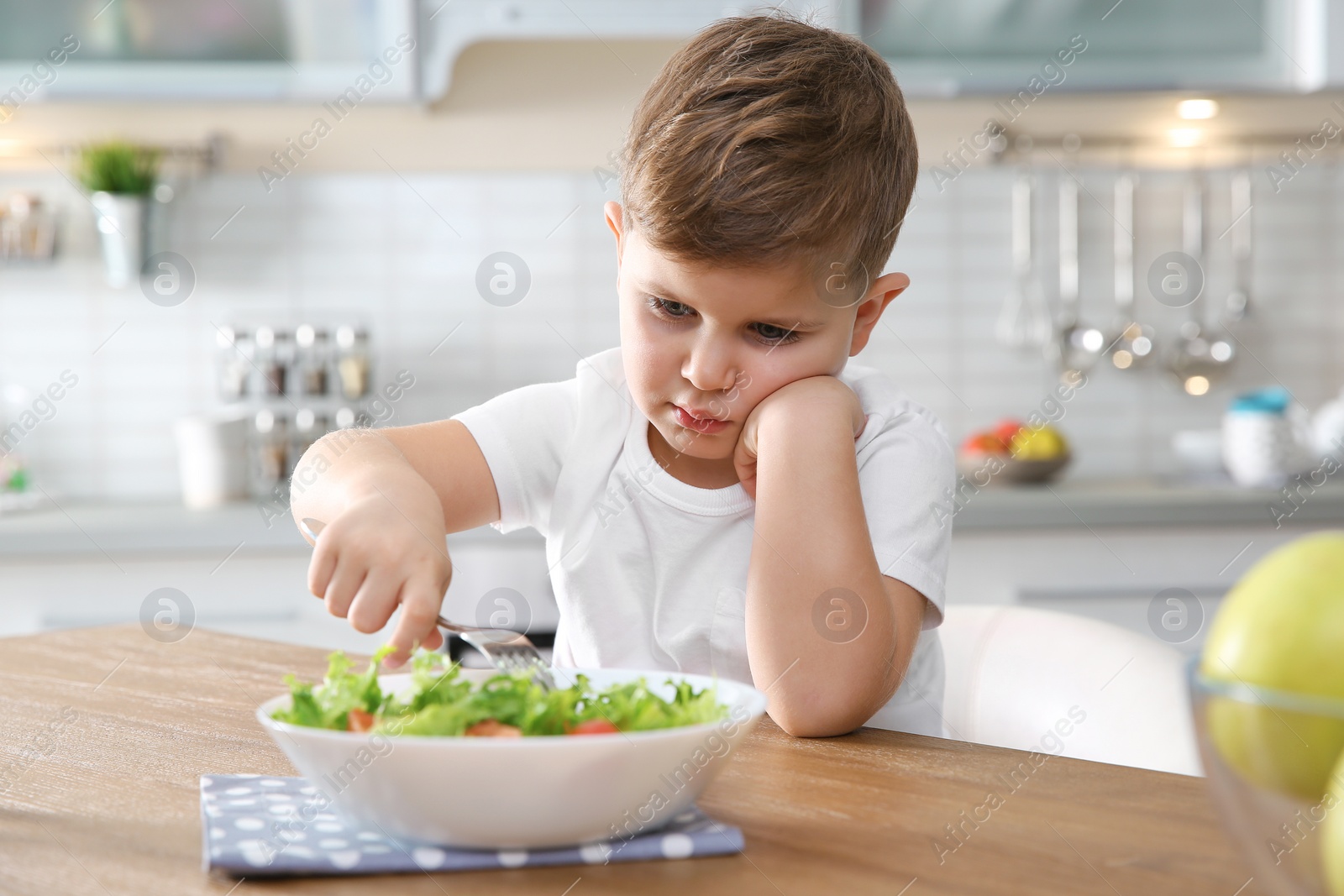 Photo of Unhappy little boy eating vegetable salad at table in kitchen