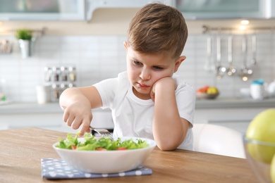 Photo of Unhappy little boy eating vegetable salad at table in kitchen