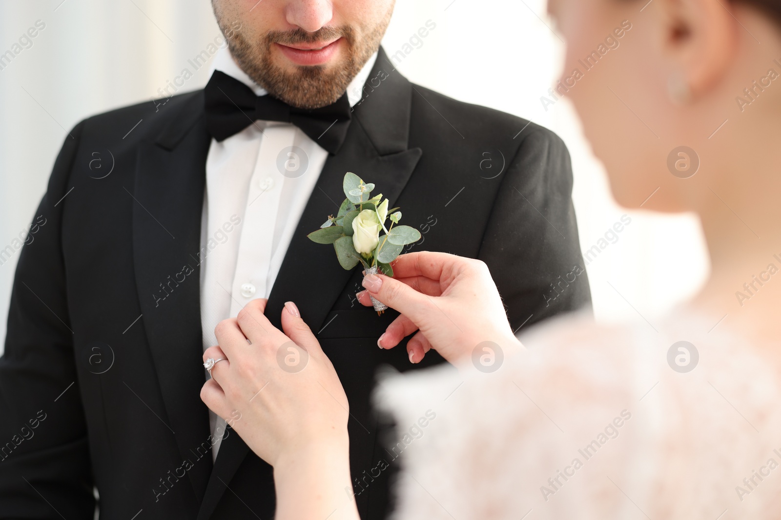 Photo of Bride putting boutonniere on her groom's jacket against light background, closeup. Wedding accessory