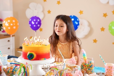 Photo of Cute little girl blowing out candles on her birthday cake indoors