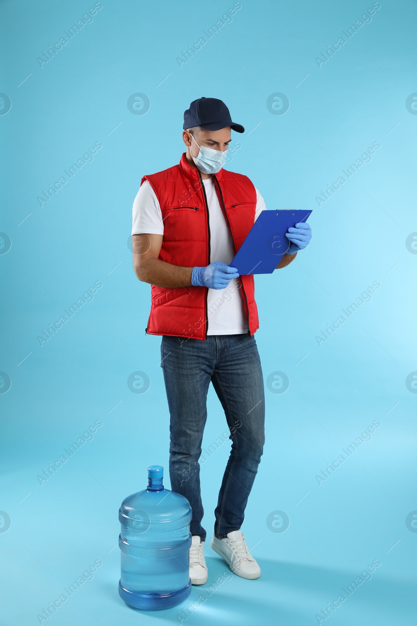 Photo of Courier in face mask with clipboard and bottle of cooler water on light blue background. Delivery during coronavirus quarantine