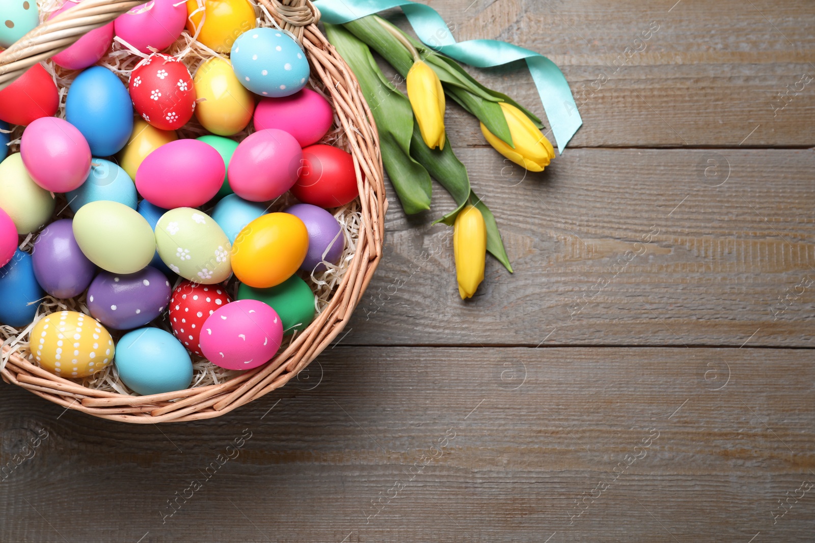 Photo of Colorful Easter eggs in wicker basket and tulips on wooden table, flat lay. Space for text