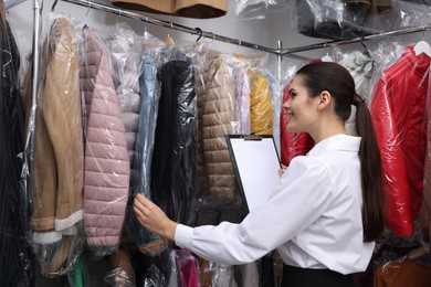 Photo of Dry-cleaning service. Happy worker with clipboard choosing clothes from rack indoors