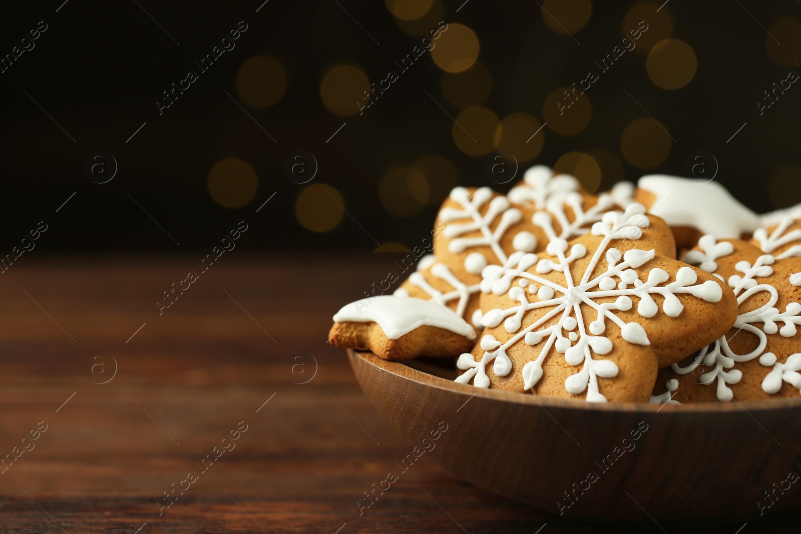 Photo of Tasty Christmas cookies with icing in bowl on wooden table against blurred lights, closeup. Space for text