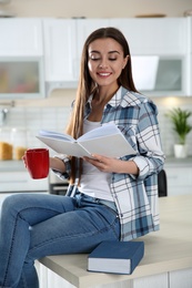 Photo of Beautiful young woman with cup of coffee reading book at home