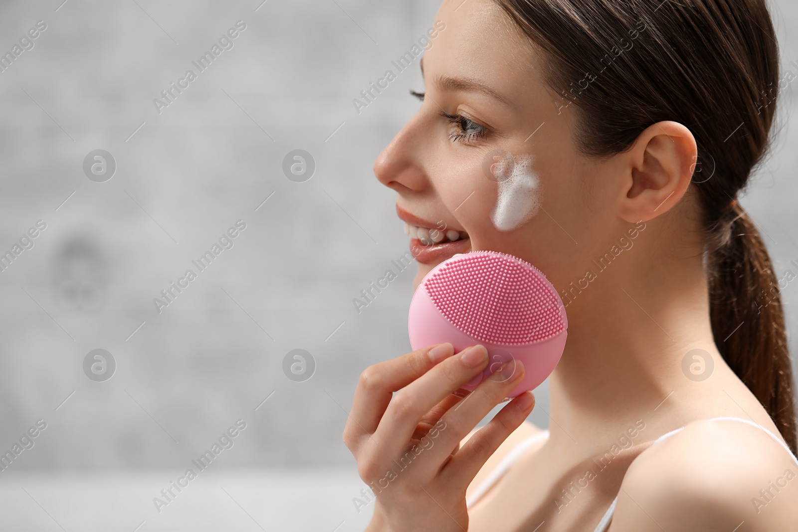 Photo of Young woman washing face with brush and cleansing foam indoors. Space for text