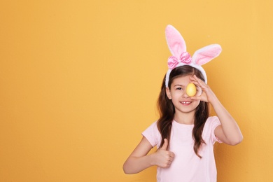 Photo of Little girl in bunny ears headband holding Easter egg near eye on color background, space for text