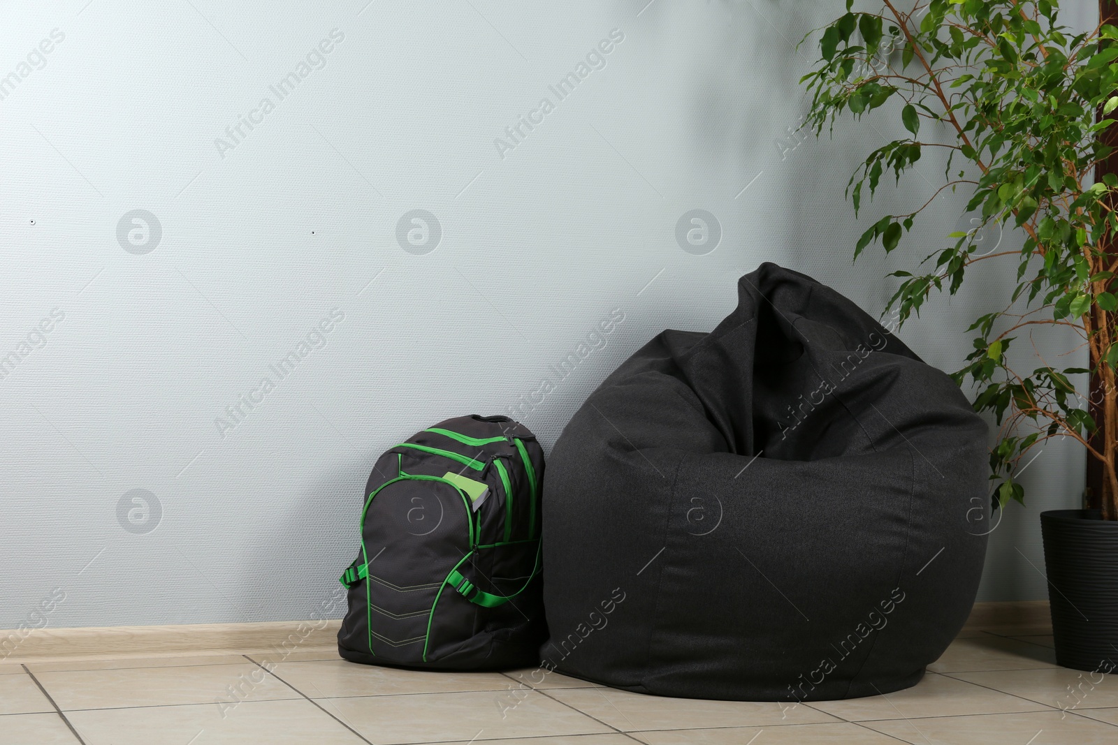 Photo of Black bean bag chair, houseplant and backpack near light grey wall in room. Space for text