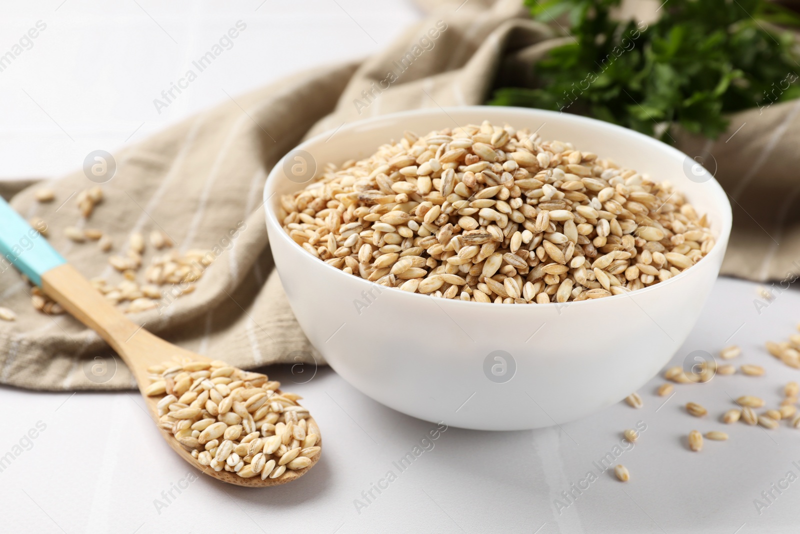 Photo of Dry pearl barley in bowl and spoon on white tiled table, closeup