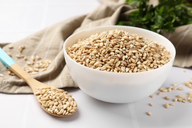 Photo of Dry pearl barley in bowl and spoon on white tiled table, closeup