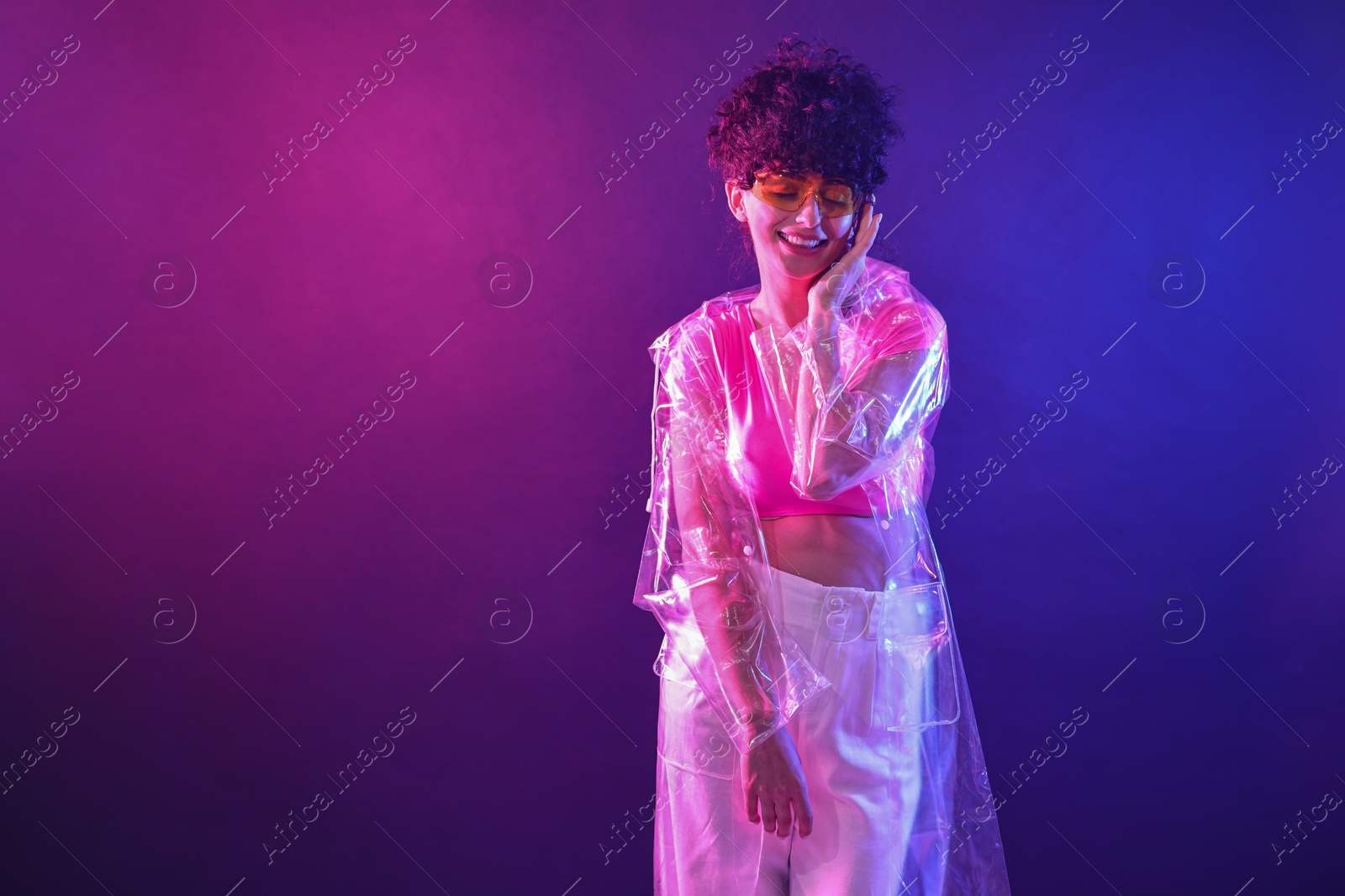 Photo of Beautiful young woman in transparent coat and sunglasses posing on color background in neon lights