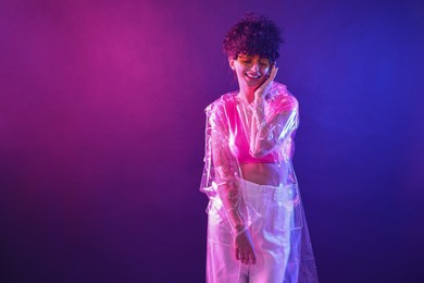 Photo of Beautiful young woman in transparent coat and sunglasses posing on color background in neon lights