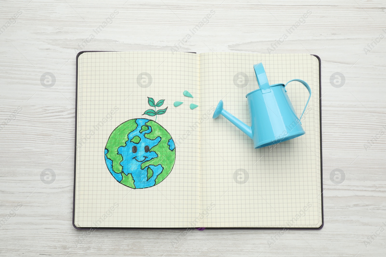 Photo of Happy Earth Day. Notebook with drawing of planet and watering can on white wooden table, top view