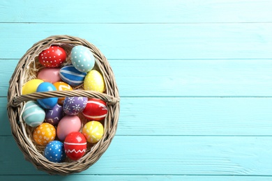 Wicker basket with painted Easter eggs on wooden background, top view. Space for text