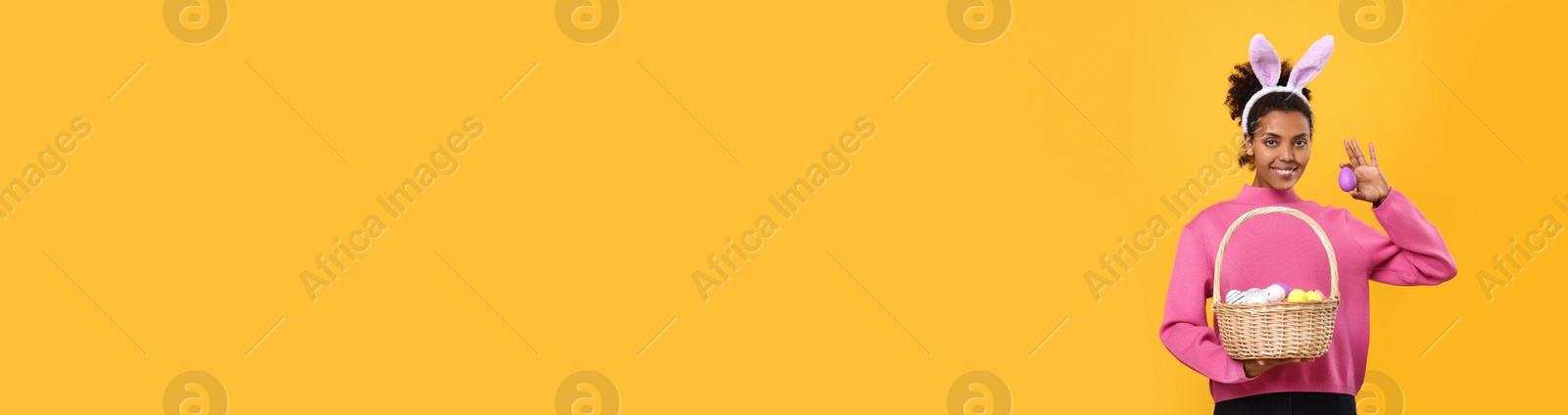 Image of African American woman with bunny ears holding basket full of Easter eggs on orange background, space for text. Banner design