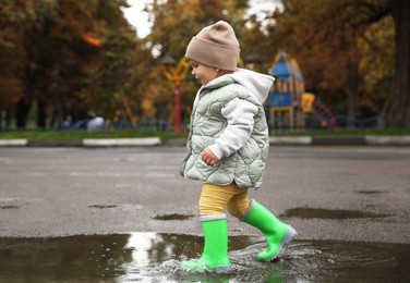 Photo of Cute little girl splashing water with her boots in puddle outdoors