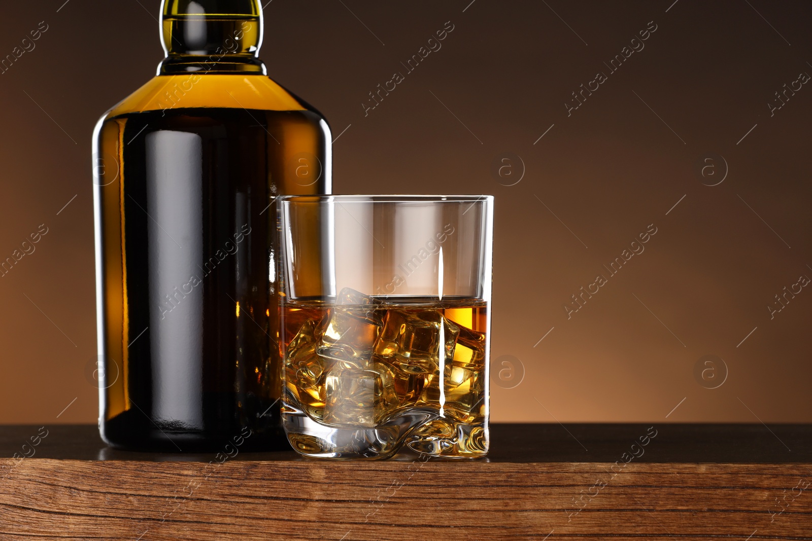 Photo of Whiskey with ice cubes in glass and bottle on wooden table, closeup. Space for text