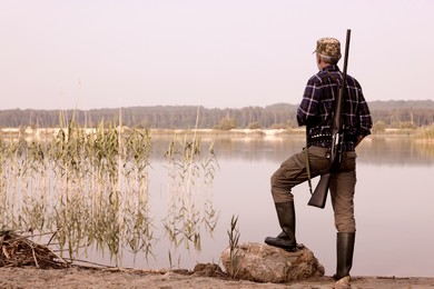 Photo of Man with hunting rifle near lake outdoors, back view. Space for text