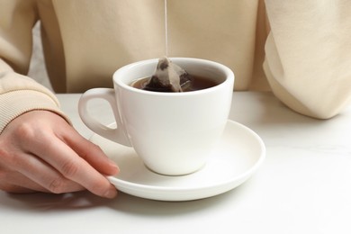 Photo of Woman putting tea bag in cup with hot water at white table, closeup