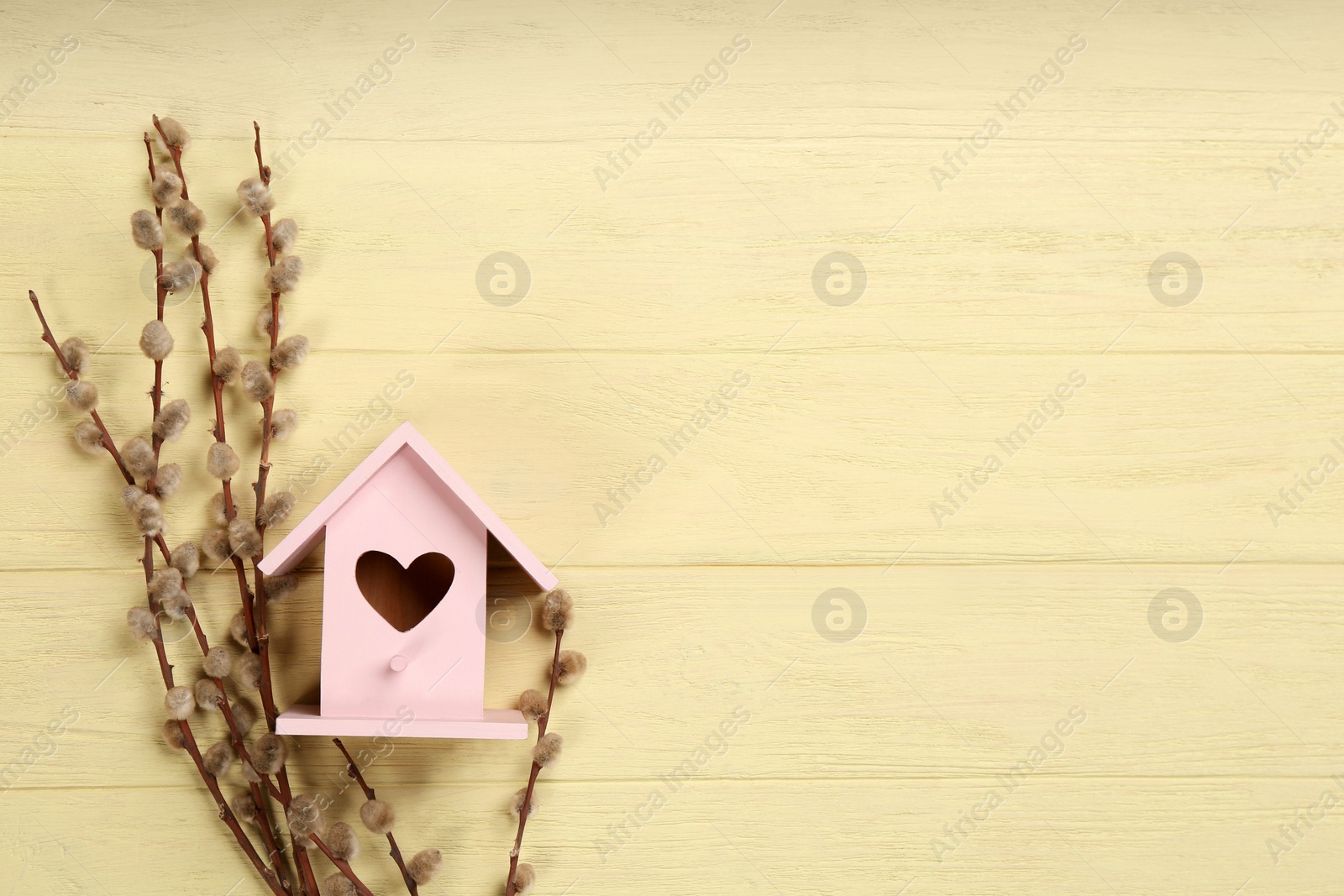 Photo of Beautiful bird house with heart shaped hole and willow branches on pale yellow wooden background, space for text. Spring flat lay composition