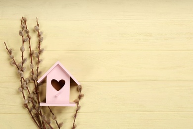 Beautiful bird house with heart shaped hole and willow branches on pale yellow wooden background, space for text. Spring flat lay composition