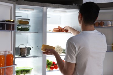 Man with cheese taking sausages out of refrigerator in kitchen at night, back view