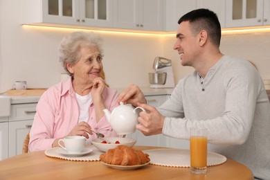 Young caregiver serving breakfast for senior woman at table in kitchen. Home care service